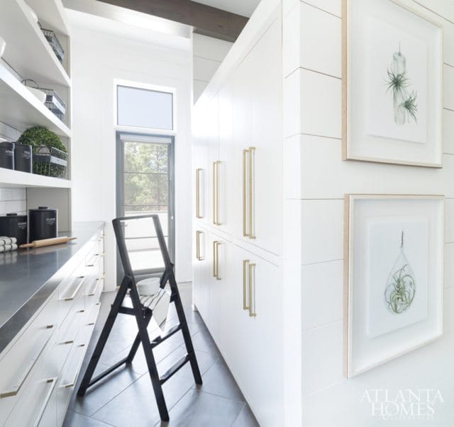 White kitchen with a long counter, white cabinetry, and tall white cabinets with brass hardware. Two framed botanical prints hang on the shiplap wall.