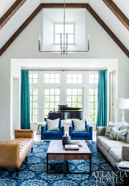 Living room with vaulted ceiling, exposed wooden beams, and a large window wall featuring a grand piano. Furnishings include blue armchairs, a tan leather bench, a grey sofa, and a patterned blue rug.