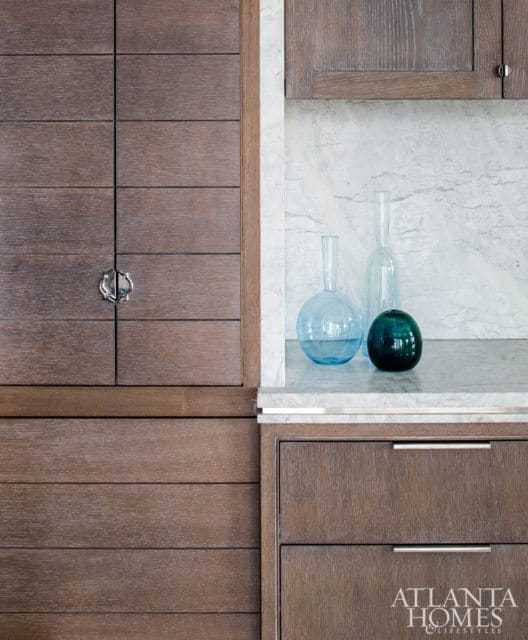 Dark wood cabinetry with horizontal plank detailing and a decorative silver pull, next to a marble backsplash and countertop holding three glass vases.