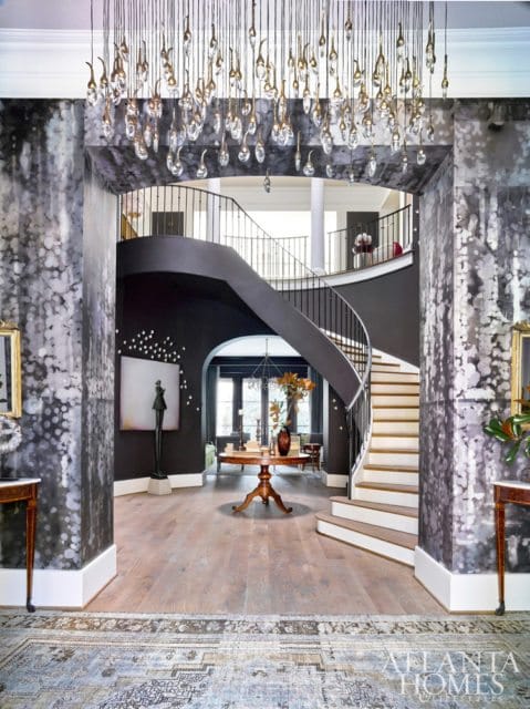 Foyer with a dramatic curved staircase, dark walls, and a large chandelier with glass teardrop accents. The walls are covered in a textured black and white wallpaper.