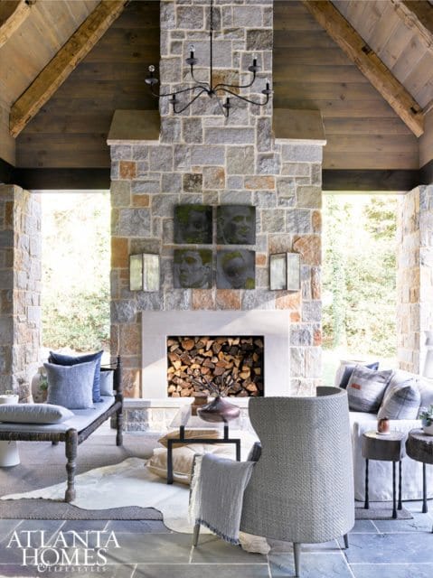 Covered outdoor living space with a large stone fireplace, wood-burning hearth, and seating area. Exposed wooden beams support the ceiling.