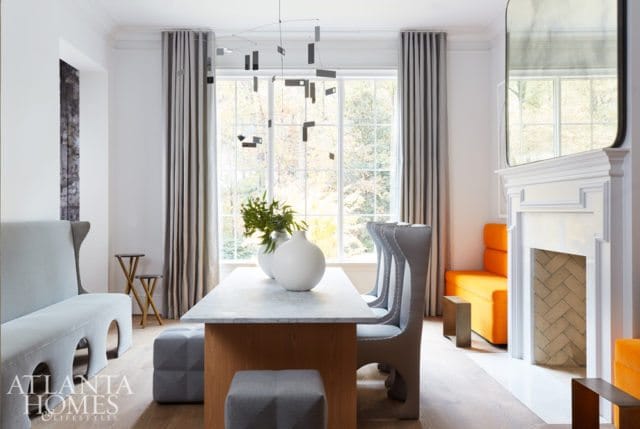 Dining room with a marble-topped table, gray upholstered chairs, a light blue bench, and a white fireplace with an orange sofa.