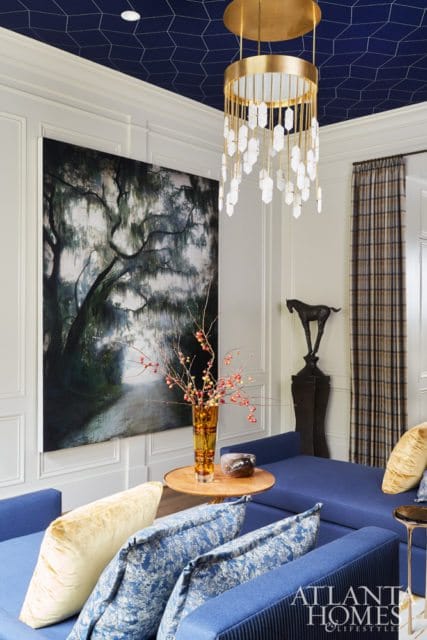 Living room with navy blue hexagonal ceiling, large artwork of a tree-lined path, blue sectional sofa with patterned pillows, and a gold chandelier with crystal accents.
