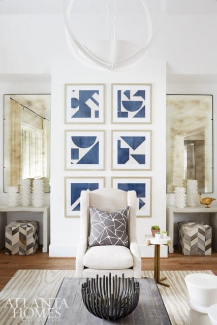 Living room with a cream upholstered armchair, a wall of six framed abstract blue and white prints, and two tall antique mirrors flanking the wall.