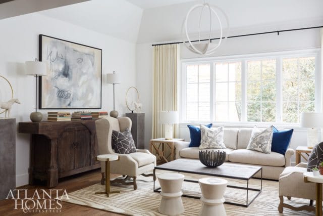 Living room with a neutral sofa, wingback chair, abstract art, dark wood buffet, and large windows.