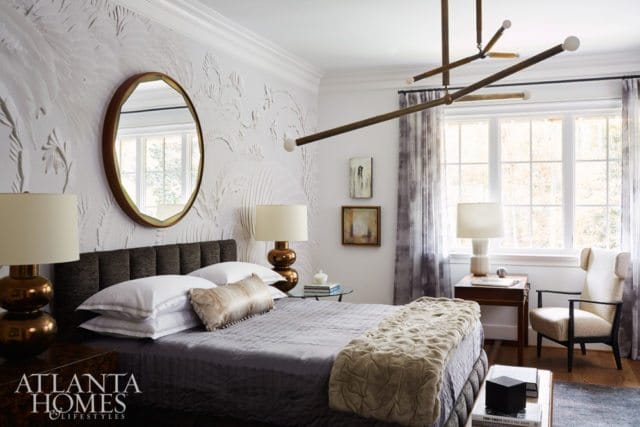 Bedroom with a textured white wallpaper, king-size bed with gray bedding, brass lamps, and a large round mirror with a wooden frame.