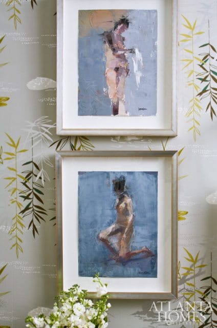 Two framed paintings of nude figures hang on a wall with botanical-themed wallpaper. The top painting depicts a standing figure, and the bottom painting shows a kneeling figure.