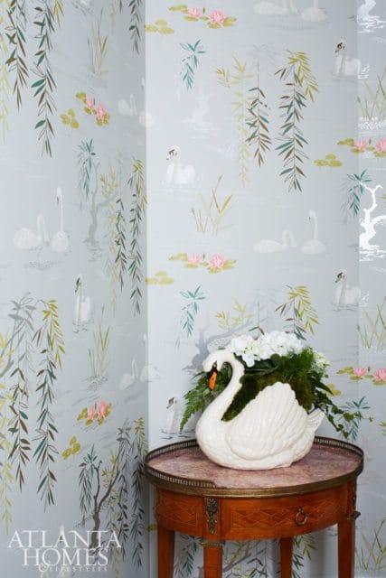 Light blue wallpaper with a repeating pattern of white swans, pink water lilies, and weeping willow branches. A white ceramic swan planter filled with white flowers sits on a small wooden table.
