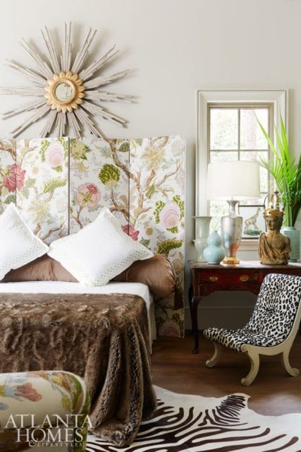 Bedroom with a floral upholstered headboard, white pillows, a brown fur throw, a sunburst mirror, and a leopard print chair next to a dark wood table with decorative objects.