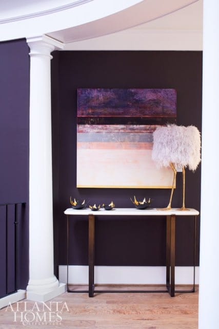 Hallway with dark purple walls, white column, console table with marble top, abstract art, and a feathered lamp.