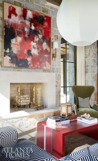 Living room with stone fireplace, abstract red and black art, green armchair, and red coffee table with books.