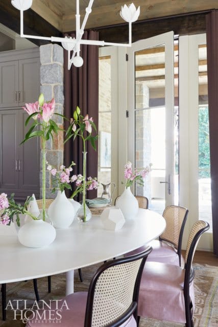 Dining room with a white oval table, cane-backed chairs with pink upholstery, and a modern white chandelier. Pink lilies in glass vases adorn the table.