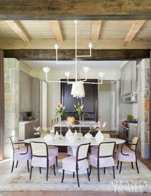Dining room with oval table, cane-backed chairs with pink upholstery, and a white chandelier. Kitchen visible in background with gray cabinetry and stone accents.