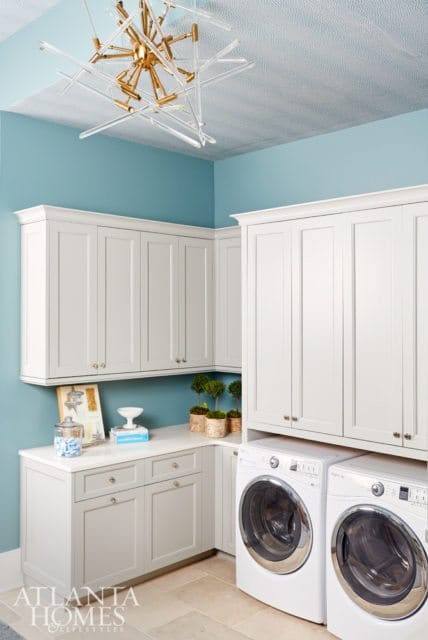 Laundry room with light blue walls, white shaker cabinets, white quartz countertops, two white front-load washers, and a modern gold and glass chandelier.