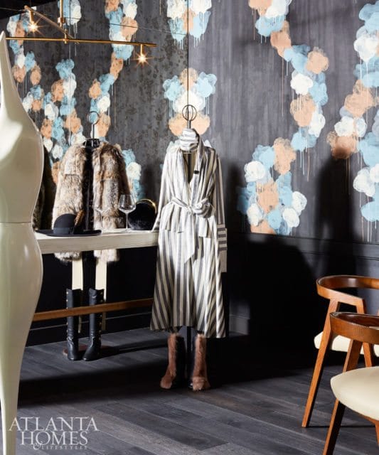 Dark wood floor in a room with a wall featuring abstract cloud-like wallpaper in blue, peach, and white. A console table displays a fur coat, hat, and wine glass, with a striped dress on a mannequin.
