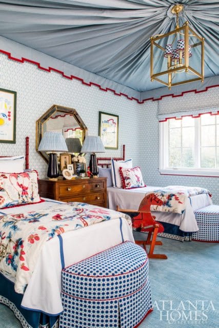 Children's bedroom with two twin beds, patterned bedding, a wooden dresser with lamps, a mirrored wall hanging, and a draped blue fabric ceiling with red trim.