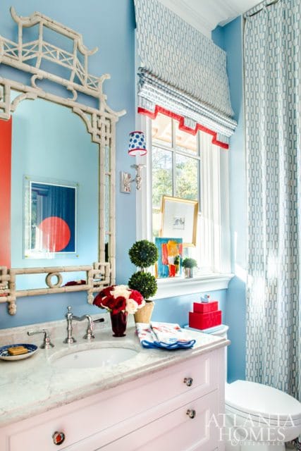 Powder room with light blue walls, marble vanity, white sink, chrome faucet, and a chinoiserie mirror. Patterned curtains and Roman shades adorn the window.