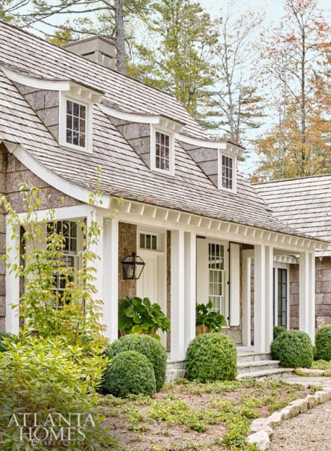 Shingle-style house with dormer windows, white columns, and a covered porch. Landscaped with manicured bushes and trees.