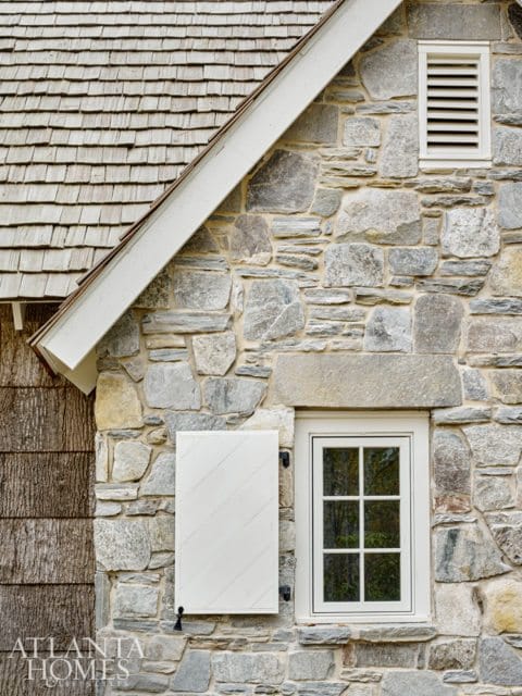 Stone exterior wall with a wooden shake roof, a louvered vent, a white shutter, and a multi-pane window.