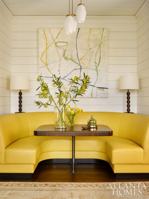 Dining nook with bright yellow curved banquette seating, dark wood table, abstract art, and two table lamps against shiplap walls.