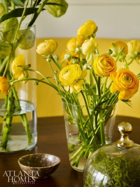 Close-up of yellow ranunculus flowers in a glass vase on a wooden table, with a small bowl and a cloche filled with moss.