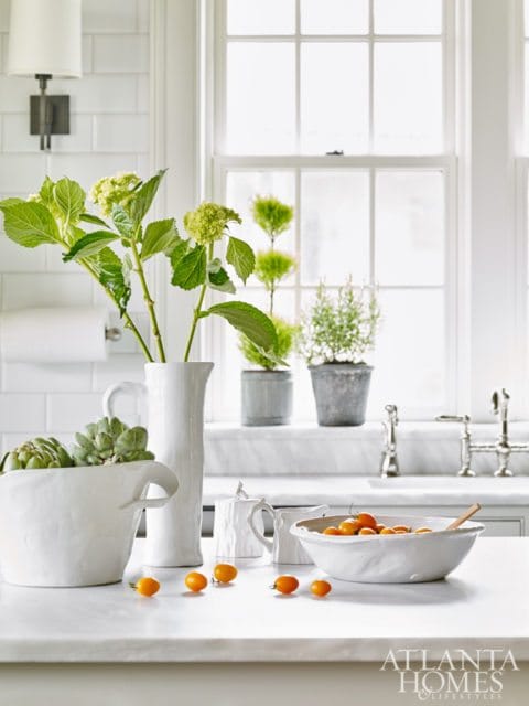 White kitchen counter with marble surface, featuring a large white textured bowl with green succulents, a tall white vase with green hydrangeas, a small white sugar pot, a white creamer, and a white bowl filled with cherry tomatoes.