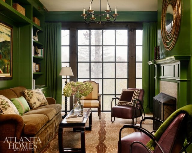 Green living room with brown sofa, leather armchairs, fireplace, and large window overlooking trees.
