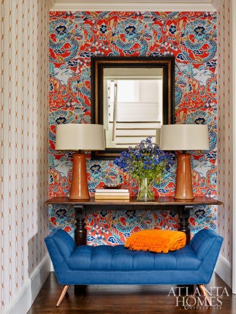 Entryway with bold red, blue, and orange dragon-patterned wallpaper, a blue tufted bench, and a console table with two orange lamps and a mirror reflecting stairs.