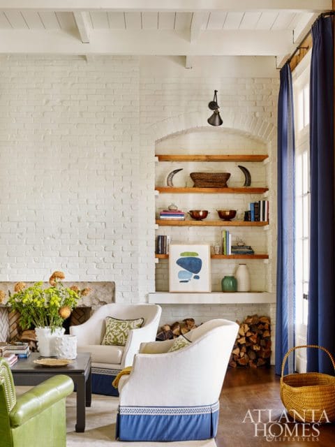 Living room with white brick walls, white armchairs with blue skirts, and built-in shelves displaying decor and books. A green leather sofa is visible in the foreground.