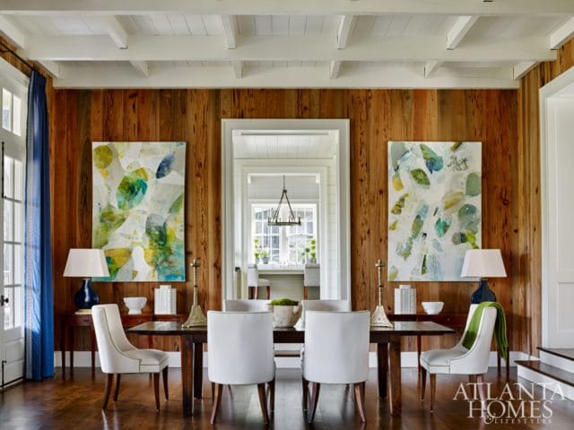 Dining room with warm wood-paneled walls, a dark wood table with white upholstered chairs, and abstract art featuring green and blue tones.