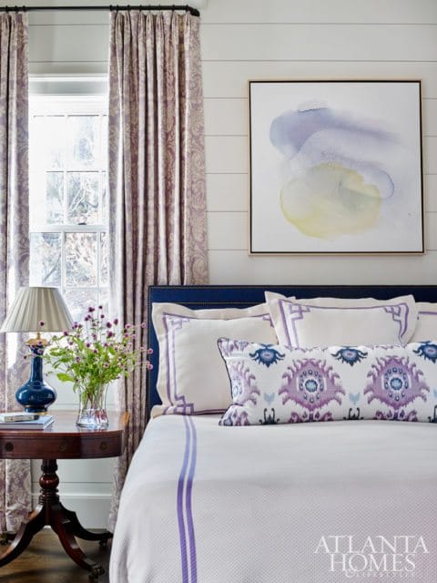 Bedroom with a navy upholstered headboard, white bedding with purple trim, patterned curtains, and abstract art on a shiplap wall.