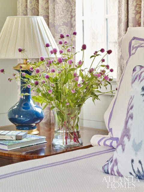 A blue ceramic lamp with a pleated cream shade sits on a wooden table next to a vase of purple globe amaranth flowers.
