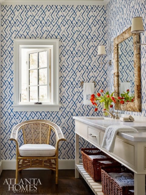 Powder room with blue and white geometric wallpaper, a rattan accent chair, a marble-topped vanity with woven baskets, and a birch-framed mirror.