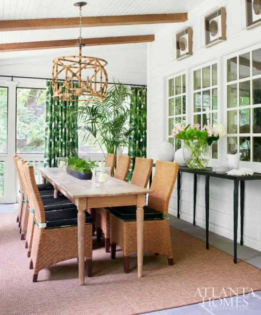 Dining room with a long wooden table surrounded by wicker chairs with black cushions, a bamboo chandelier, and green floral curtains.