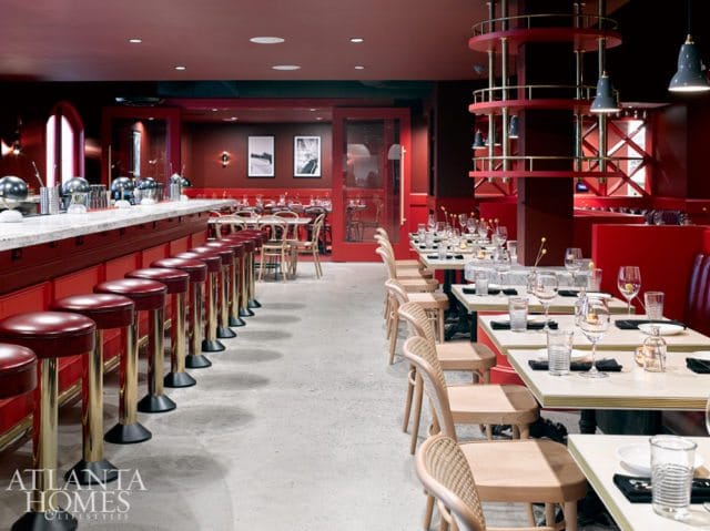 Restaurant interior with red banquettes, a long marble-topped bar with red stools, and tables set with glassware. Gold accents and pendant lights are visible.