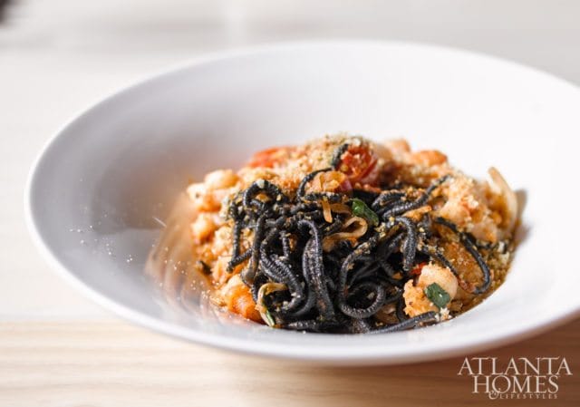Black squid ink pasta with shrimp and cherry tomatoes, topped with breadcrumbs, served in a white bowl with the Atlanta Homes & Lifestyles logo in the corner.