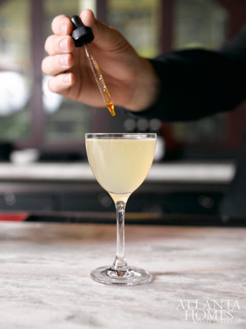 Close-up of a hand holding a dropper with amber liquid, adding a drop to a pale yellow cocktail in a coupe glass.