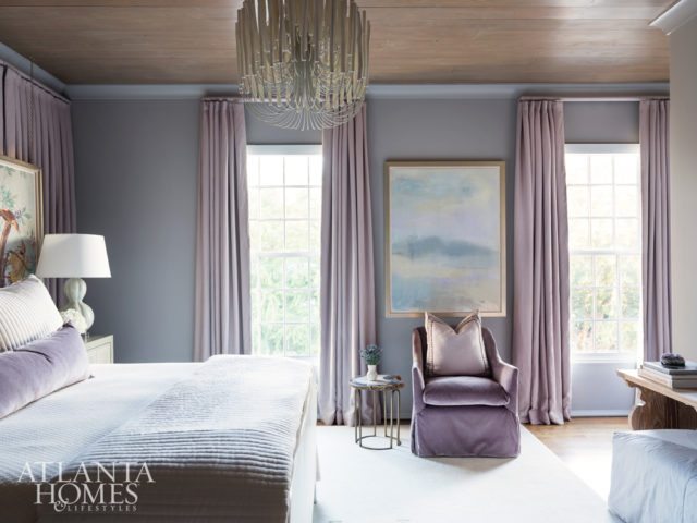 In this guest suite which is painted Sherwin-Williams Proper Gray, a wall of drapery panels hides unusable windows and absorbs noise to create a calming retreat. In front of the panels, artwork is suspended from the ceiling by brass chains. Purple hues abound in a plush chair from LEE Industries and drapery fabric through Pindler. The chandelier is through Arteriors.