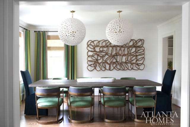 Dining room with a dark wood table, green upholstered chairs with brass frames, and navy slipcovered chairs. Two large white orb chandeliers hang above.