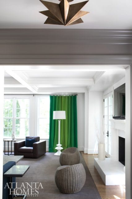 Living room with coffered ceiling, brown leather armchair, green ombre curtains, white floor lamp, and two woven ottomans.
