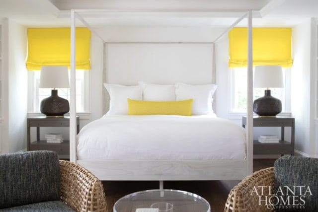 Bedroom with a white four-poster bed, white bedding, and a yellow lumbar pillow. Yellow Roman shades hang above nightstands with dark lamps.
