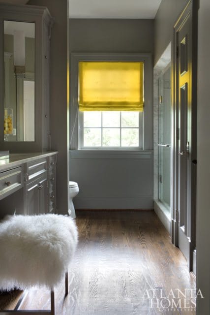 Gray bathroom with a vanity, white fluffy stool, toilet, glass shower enclosure, and a bright yellow Roman shade over a window.