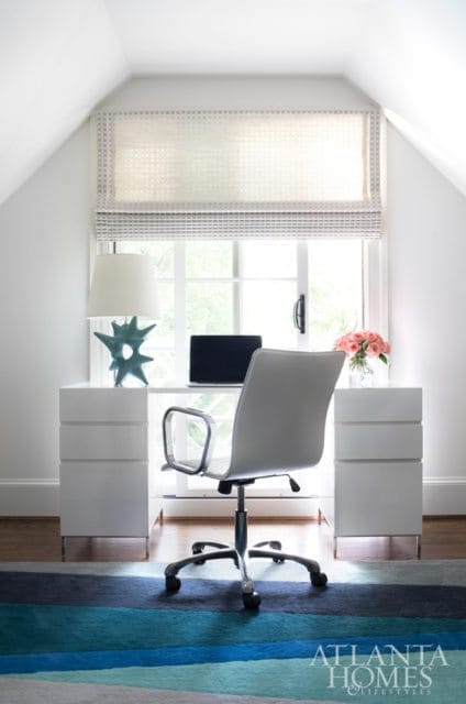 White desk with a laptop and a blue sculptural lamp, facing a window with a Roman shade. A grey office chair is in front of the desk, on a striped rug.