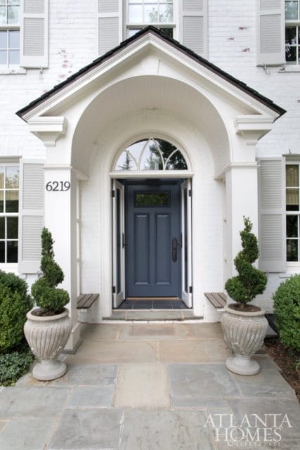 White brick house with gray shutters and a dark blue arched entryway door. Flanked by potted topiaries and stone walkway.