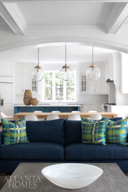 Navy blue sofa with plaid pillows in front of a long wooden dining table and white chairs, with a white kitchen and island in the background.