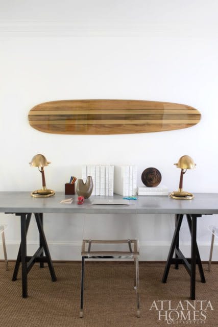 Modern home office with a metal desk, two brass desk lamps, a laptop, and a vintage wooden surfboard mounted on a white wall.