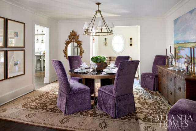 Dining room with plush purple velvet chairs around a round table, ornate gold mirror, and a landscape painting above a wooden buffet.