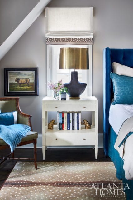 Bedroom with a blue tufted headboard, a green armchair with a blue throw, a white nightstand with books and a lamp, and a fawn-print rug.