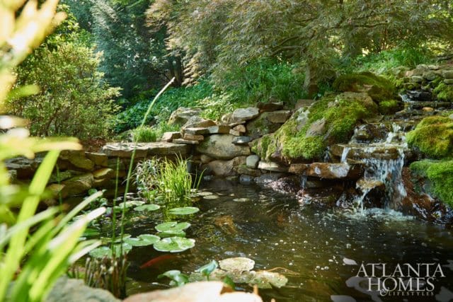 Pond with lily pads and koi fish, surrounded by moss-covered rocks and a small waterfall, with lush greenery and trees in the background.