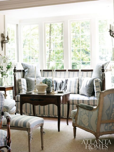 Living room with striped sofa, patterned armchairs, and a wooden side table in front of large windows overlooking trees.